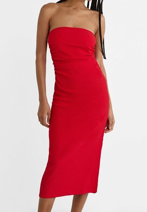 Vrouw draagt een strapless, aansluitende rode midi-jurk, staand tegen een effen lichte achtergrond.