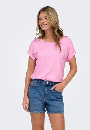 Donna che indossa una maglietta rosa a maniche corte infilata in pantaloncini di jeans blu, in piedi con una mano in tasca contro uno sfondo semplice.