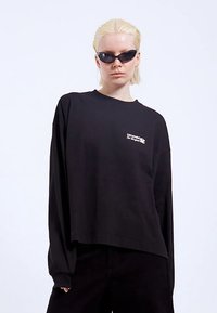 Černé tričko s dlouhým rukávem v uvolněném střihu, s bílým grafickým logem na hrudi. Hladká bavlněná látka a spadlými rameny.