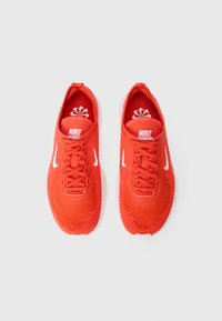 Piros Nike sportcipők, hálós külsővel, kerek orral, fehér logókkal és texturált fűzőkkel. Párnázott talpbetét, amely virágmintás.