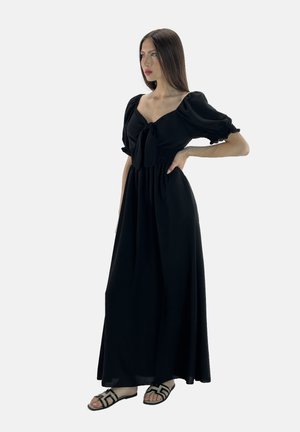 Robe maxi noire avec manches bouffantes, décolleté à nouer, buste texturé et jupe fluide. Portée avec des sandales plates noires dotées d'acents géométriques.