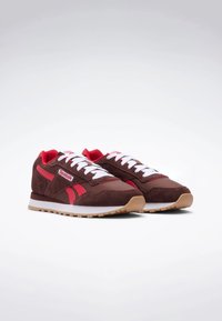 Braune und rote Sneaker mit einem Obermaterial aus Wildleder, weißen Schnürsenkeln und einer hellbraunen Gummisohle. Mit einem auffälligen Reebok-Logo an der Seite.