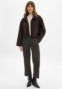 Bruine cropped jack met oversized zakken, gecombineerd met broek in houndstooth patroon en zwarte haklaarzen. Lichtbeige top eronder.