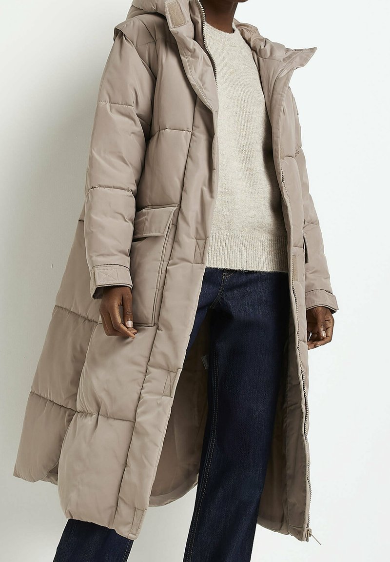 Abrigo acolchado largo y beige con capucha, que presenta un cierre de cremallera, bolsillos laterales y un diseño acolchado, combinado con un suéter ligero y jeans.