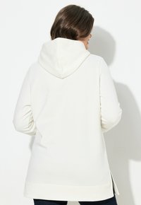 Sudadera blanca con capucha, de corte relajado, mangas largas y aberturas laterales. Textura suave, diseño minimalista y detalles en la cintura.