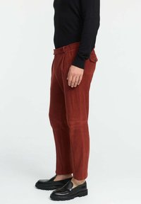 Rostfarbene Cordhosen mit geradem Schnitt, ausgestattet mit einem Gürtel, Seitentaschen und einer einzelnen Gesäßtasche. Kombiniert mit schwarzen Slip-On-Schuhen.