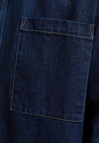 Tessuto in denim blu scuro con una tasca a toppa. Presenta cuciture visibili di un colore beige a contrasto, che mettono in risalto i bordi e le cuciture della tasca.
