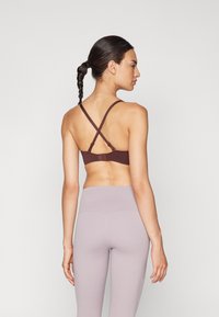 Καφέ αθλητικό σουτιέν με σταυρωτές μπούστες στην πλάτη, συνδυασμένο με ψηλόμεσες ανοιχτό μωβ κολάν. Το υλικό φαίνεται λείο και ελαστικό.