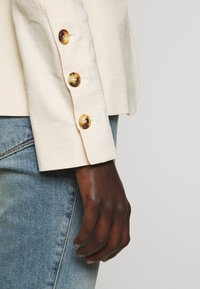 Krémový lněný blazer s rukávy a čtyřmi velkými želvovinovými knoflíky. Na manžetě je viditelná světle modrá džínová látka s mírnou texturou.