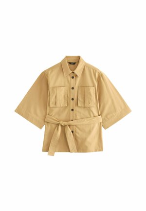 Beige kortermet skjorte med frontknapper, to brystlommer og en bindebælte i taljen på en hvid baggrund.
