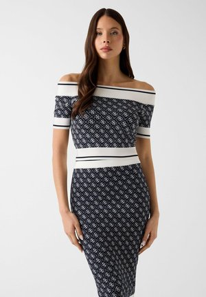 Donna con capelli castani lunghi che indossa un abito aderente blu navy con spalle scoperte, motivo geometrico bianco e bordino bianco su maniche e vita.