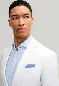 Weißer Blazer mit Revers über einem hellblauen Leinenhemd. Ein gemusterter blauer Einstecktuch akzentuiert die Tasche.