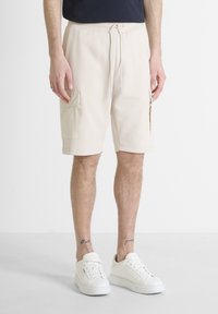 Pantalones cortos de cargo beige con cinturilla elástica, cordón y dos bolsillos laterales. Combinados con zapatillas blancas sobre un fondo claro.