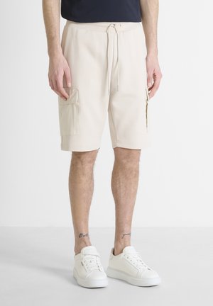 Beige cargoshorts met elastische tailleband, trekkoord en twee zijzakken. Gecombineerd met witte sneakers op een lichte achtergrond.