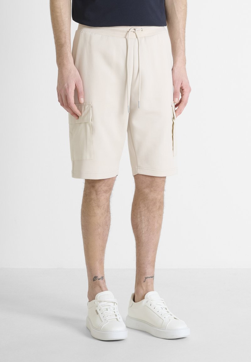Pantalones cortos de cargo beige con cinturilla elástica, cordón y dos bolsillos laterales. Combinados con zapatillas blancas sobre un fondo claro.