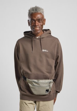 Bruine hoodie met een voorzak en logo. Grijze heuptas die om de taille is gedragen, met een rits en merketiket. Beige broek.