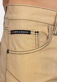 Pantalons en denim beige avec des coutures noires contrastantes, une petite étiquette noire et rouge sur la poche arrière, et une texture lisse.