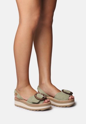 Noge v zelenih semiš sandalah na pladnju z velikim okroglim zaponkam in nastavljivimi zanke na svetli podlagi.