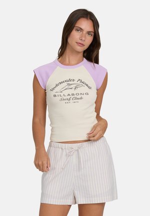 Kremowy crop top z lawendowymi krótkimi rękawami, na których znajduje się nadrukowane logo. Połączony z jasnymi pasiastymi szortami z wiązanym w pasie sznurkiem.