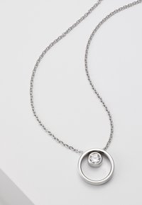Collier en chaîne argentée avec un pendentif circulaire orné d'une gemme claire insérée dans un anneau plus grand. Texture lisse, design moderne.