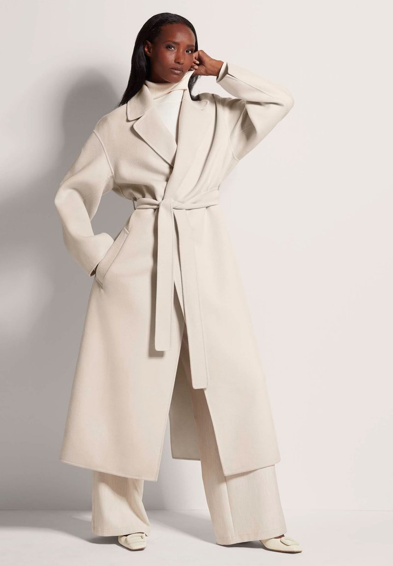 Lange beige wikkeljas met brede mouwen, bijpassende riem en zijzakken, gelaagd over een geribde witte coltrui en brede beige broek.