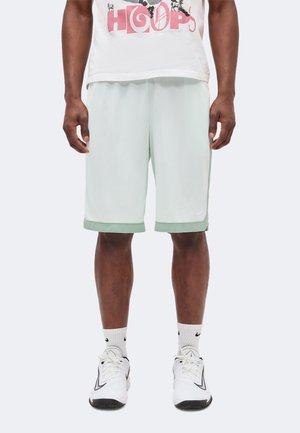 Mann trägt weiße Basketball-Netzshorts mit hellgrünem Rand, weiße Nike-Socken und weiße Nike-Sneakers und steht vor einem einfarbigen Hintergrund.