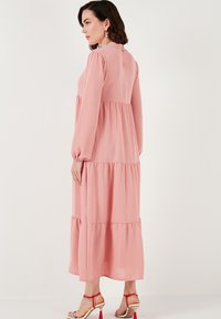 Robe longue rose avec trois niveaux, manches longues et col rond. Fabriquée en tissu doux ; présente de légers détails froncés aux épaules.