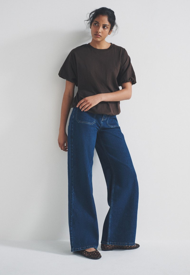 Ung kvinde står op ad hvid væg iført sort kortærmet top, vidbenede mørkeblå jeans og sorte mønstrede flade sko.