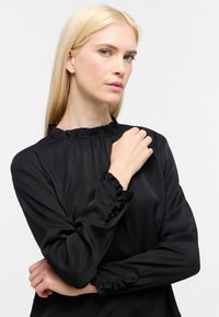 Blusa nera a maniche lunghe con collo e polsini arricciati, realizzata in tessuto liscio, caratterizzata da una vestibilità comoda e un design semplice.