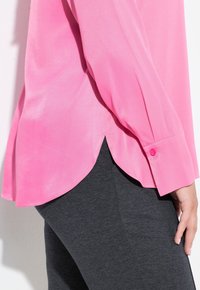 Lyserød bluse med lange ærmer i silkeagtig tekstur og buet nederste kant, kombineret med mørkegrå bukser. Knappetalje på ærmekanten.