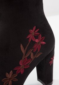 Stivale alto in suede nero con design floreale ricamato in rosso scuro e marrone lungo il lato e il tacco.