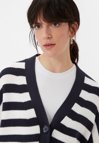 Marineblau und weiß gestreifter Cardigan mit V-Ausschnitt, Knopfschluss und strukturiertem Strickstoff, über einem weißen Rippoberteil getragen.