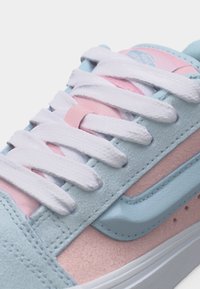 Κοντινό πλάνο κορδονιών παπουτσιού σε σουέντ παστέλ μπλε και ροζ, με λευκά κορδόνια και εμφανές μπλε λογότυπο σβουριάς.