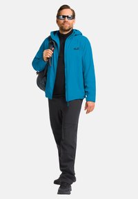 Giacca leggera blu con cappuccio, indossata sopra un top in pile nero e pantaloni neri. L'uomo tiene uno zaino e indossa occhiali da sole.