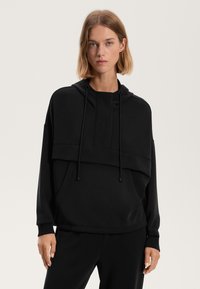 Felpa nera con cappuccio realizzata in tessuto morbido, con colletto alto, coulisse, una grande tasca frontale e un design dal taglio rilassato con maniche lunghe.