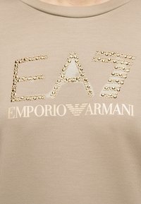 Μπεζ ύφασμα με χρυσά στρας που σχηματίζουν το "EA7" πάνω από το τυπωμένο κείμενο "Emporio Armani" και το λευκό λογότυπο αετού της μάρκας.