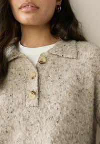 Bežový pletený sveter s textúrovaným povrchom, s golierom a tromi veľkými, svetlými gombíkmi. Nosený cez biely top.