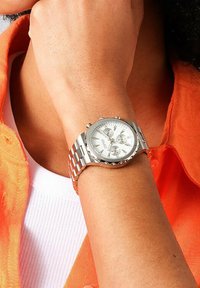 Silverfärgad rostfri stålarmband med rund urtavla, vit urtavla, tre subur och en länkad armband. Bärs på ett handled mot ett orange tyg.