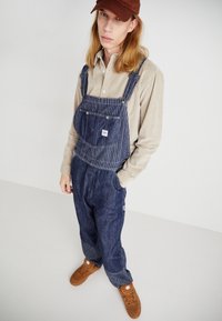 Pantalones de mezclilla con sección superior a rayas, pantalones azul oscuro, camisa de manga larga crema debajo y zapatillas marrones. Una gorra de visera plana completa el look.