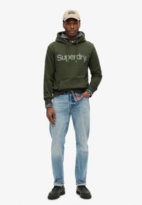 Oliven grøn hættejakke med "Superdry" logo, lagdelt over en ternet skjorte. Bærende med lyseblå jeans og sorte sko. Beige kasket fuldender looket.