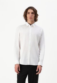 Cămașă albă cu mânecă lungă, din țesătură de in, cu guler clasic și croială dreptă, asortată cu pantaloni negri.