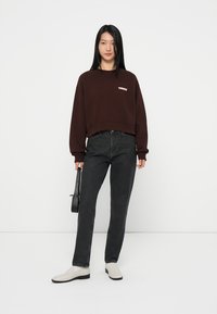 Felpa corta marrone con logo, abbinata a jeans neri a vita alta e scarpe off-white. Il modello tiene in mano una piccola borsa nera. Sfondo semplice.