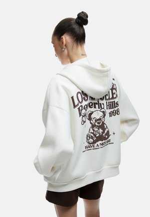 Hvid oversized hættetrøje med et grafisk print af en tøjdyr og tekst på ryggen. Har snore i hætten og dropped shoulders.