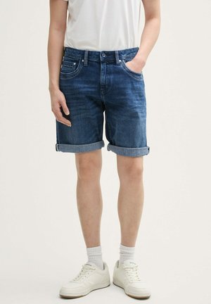 Dunkelblaue Jeansshorts mit umgeschlagenen Säumen, ausgestattet mit einem klassischen Fünf-Taschen-Design und subtilen Verblassungen. Kombiniert mit weißen Sneakers.