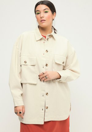 Beige overshirt van textiel met voorkruiszakken en bruine knopen. Lange mouwen, losse pasvorm en afgeronde onderkant.