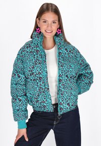 Jachetă puf turquoise cu imprimeu leopard negru și roz, guler înalt, închidere cu fermoar și mansete contrastante. Modelul poartă un top alb și blugi.