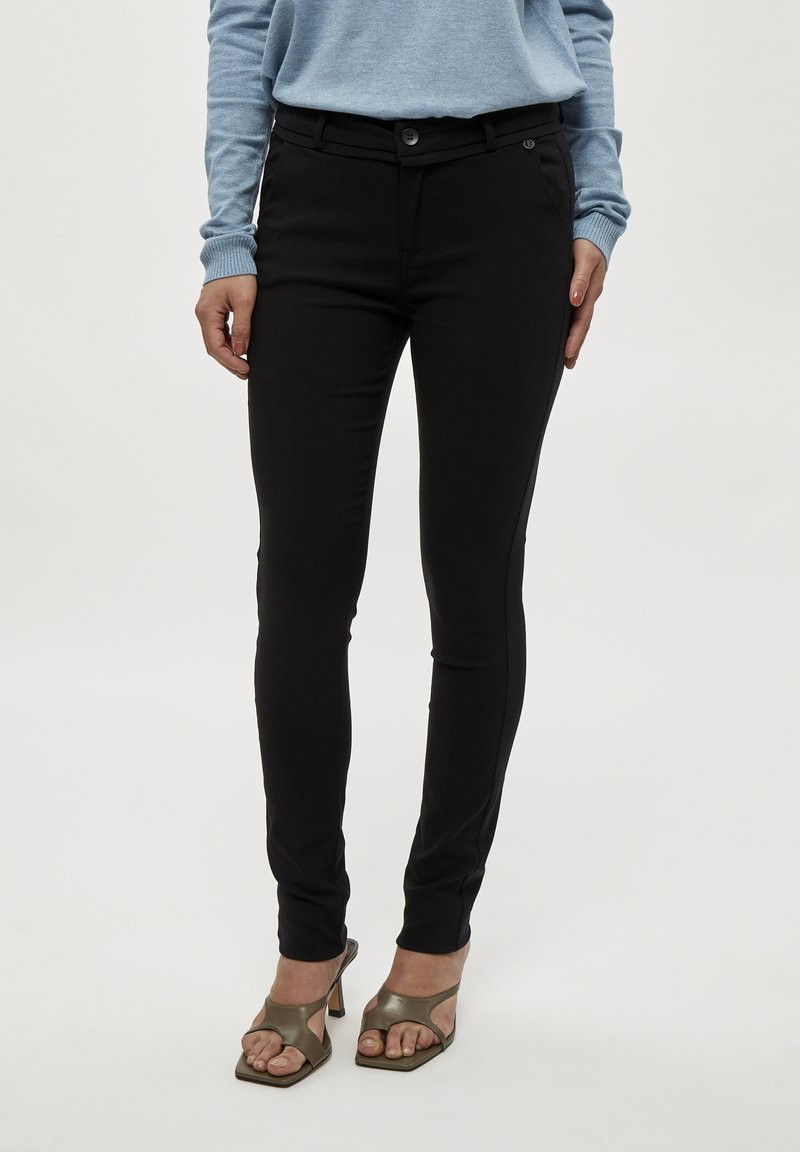 Zwarte skinny broek met een gladde textuur, voorzien van een knoopsluiting en een voorzak. Draagt samen met olijfgroene open teen hakken.