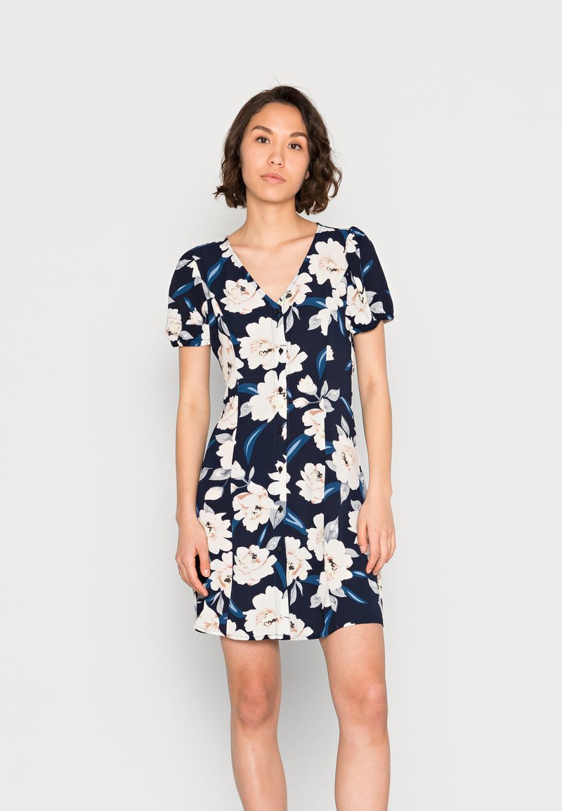 Vestido floral com fundo azul- Escuro, grandes flores brancas e folhas verdes, decote em V e mangas curtas bufantes.