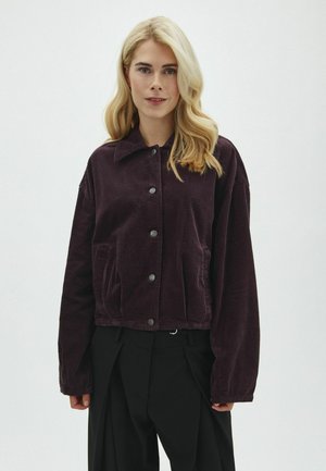 Femme blonde portant une veste en velours côtelé violet foncé boutonnée et un pantalon noir ample sur un fond clair uni.
