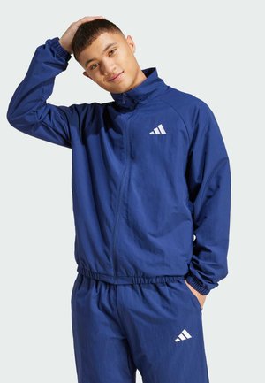 Marineblå zip-jakke lavet af letvægtsmateriale, med elastiske manchetter og talje, samt et hvidt logo på brystet.